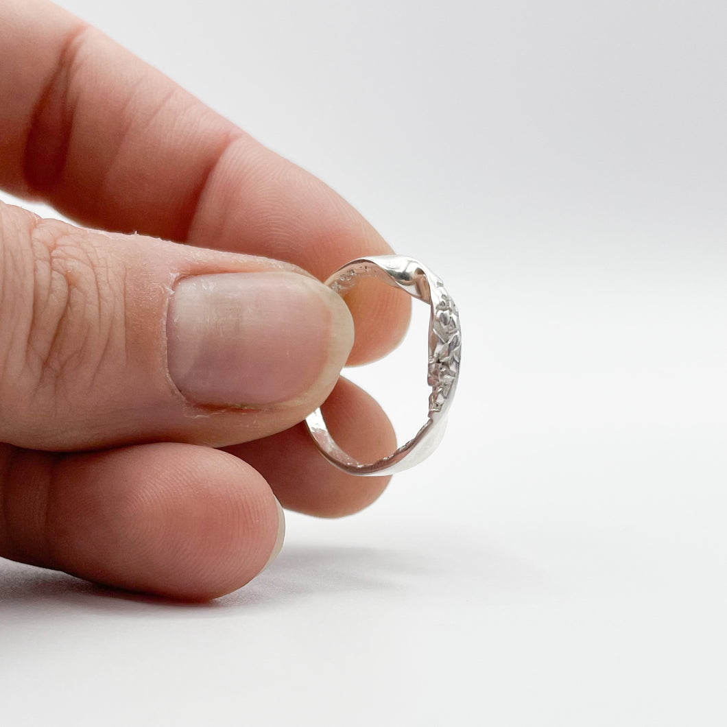 Silver ring held in fingertips, showing a 3/4 view, showing the top third of the ring is twisted, with the 'inside' edge showing a chipped, rocky texture. 