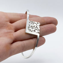Load image into Gallery viewer, A round cuff bracelet sprouts from either side of a square of metal with a raised &#39;bubble&#39; texture- shown draped over fingers.
