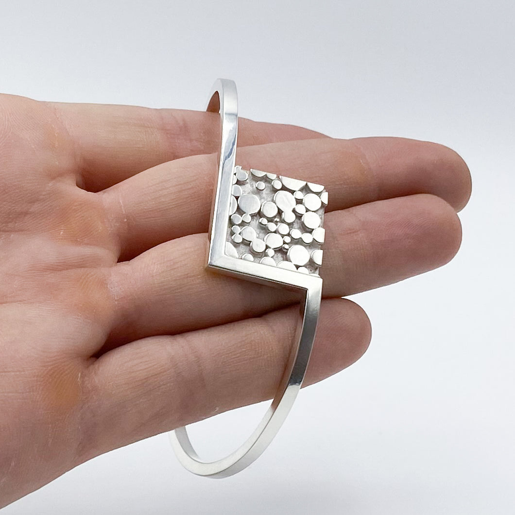 A round cuff bracelet sprouts from either side of a square of metal with a raised 'bubble' texture- shown draped over fingers.